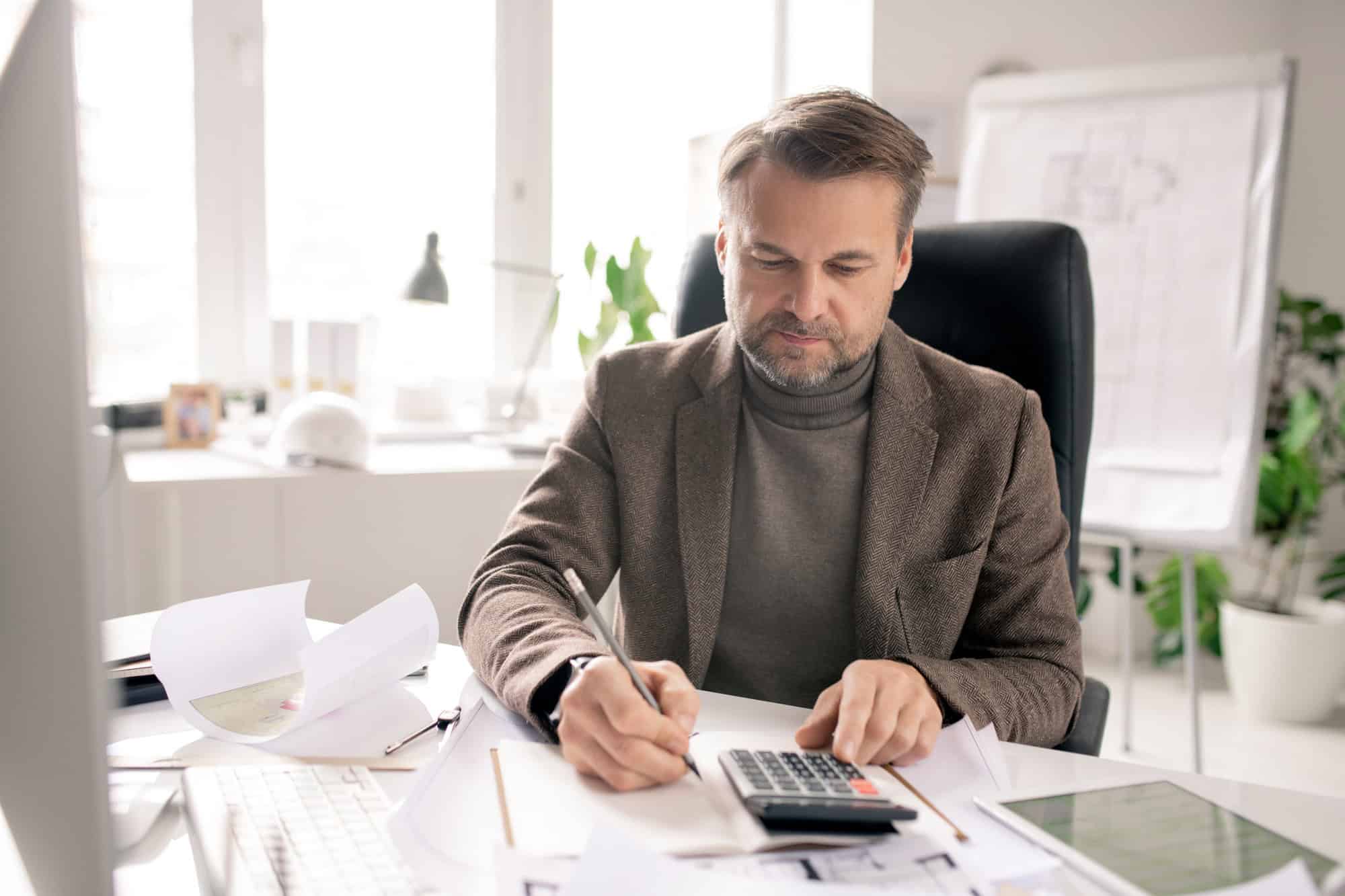Mature engineer or accountant with pen and calculator making notes in notebook